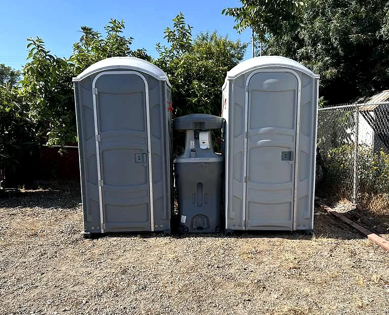 Two grey standard portable toilets with a portable handwashing station between them, set up on a dirt lot under a clear sky