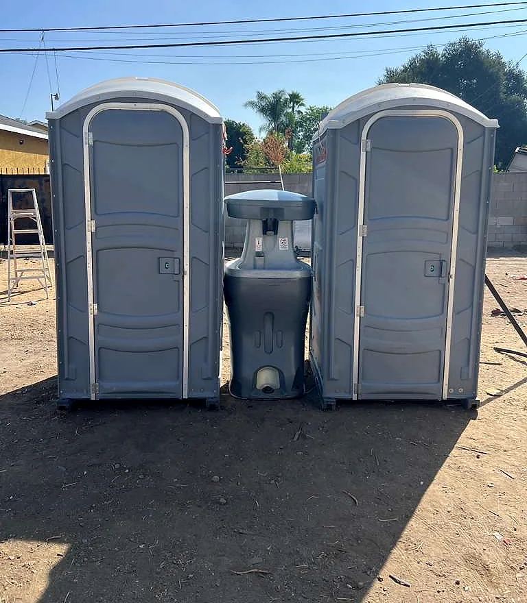 Two standard portable toilets flanking a portable handwashing station on a graded dirt jobsite