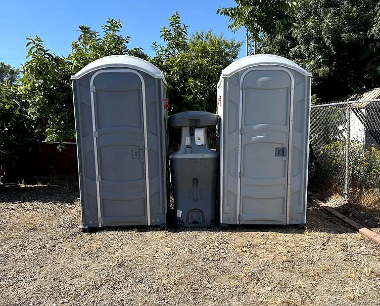 Two standard grey portable toilets with a handwashing station between them on a gravel lot